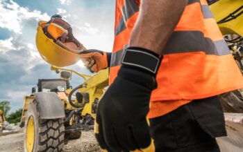 Trabalhador em obra segurando capacete de segurança próximo a maquinário e gerador de energia para obras.