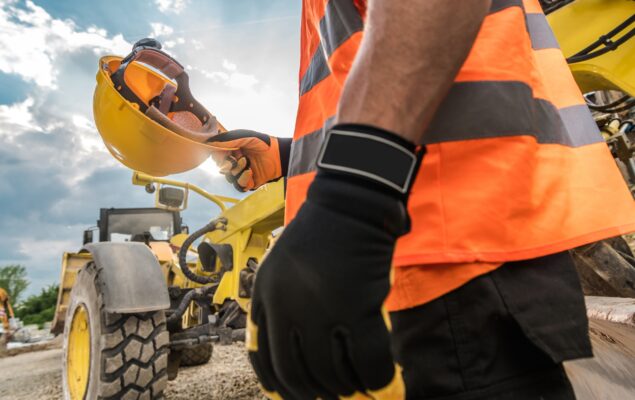 Trabalhador em obra segurando capacete de segurança próximo a maquinário e gerador de energia para obras.